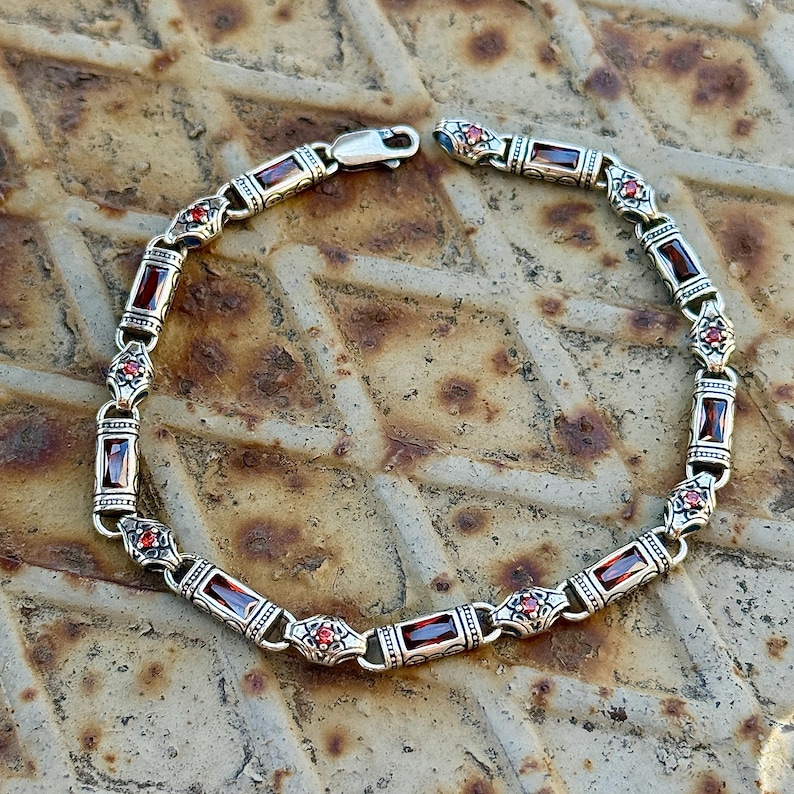 May include: Silver bracelet with rectangular red gemstones and small red accent stones. The bracelet has a decorative design with a clasp closure. The jewelry is displayed on a textured, metallic surface.