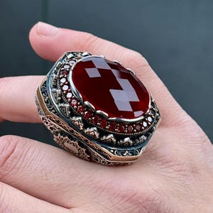 Puede incluir: Un anillo ornamentado con una gran piedra preciosa roja oscura tallada en forma ovalada. El anillo tiene una banda plateada detallada con detalles dorados y piedras rojas más pequeñas. El anillo está en una mano. El diseño es intrincado.