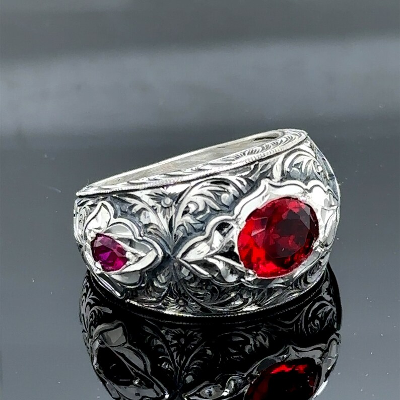 Peut inclure: Une bague en argent avec des motifs floraux complexes et deux pierres pr&eacute;cieuses rouges. La bague est large et de forme arrondie.