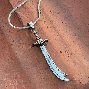 Puede incluir: Un collar de plata con un colgante de espada de plata detallada. La espada tiene una hoja curva con diseños intrincados y un mango de color dorado.