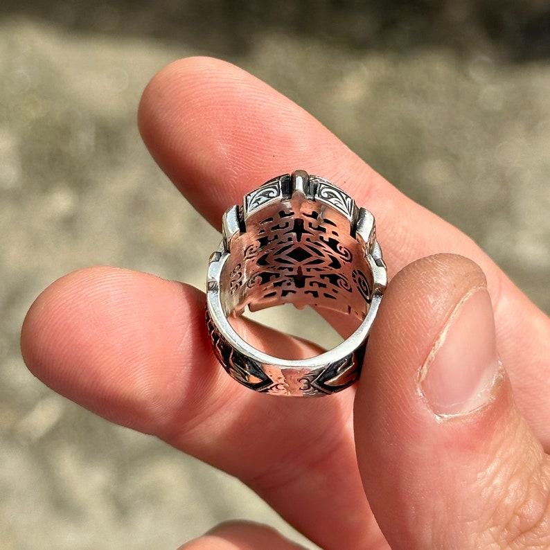 May include: A silver ring with an intricate, detailed design. The ring is worn on a finger and the design is visible on the top and sides of the ring.