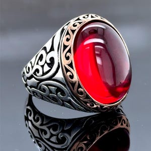 Könnte beinhalten: Ein silberner Ring mit einem roten Edelstein, der in einem dekorativen Filigranmuster gefasst ist. Der Ring hat einen goldfarbenen Akzent um den Stein.