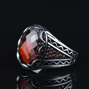 Pode incluir: Um anel de prata com uma pedra preciosa vermelha oval e facetada. O anel tem detalhes intrincados ao redor da pedra preciosa e ao longo da banda, com pedras vermelhas menores acentuando o design. O anel é exibido em um fundo preto.