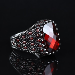 May include: A silver-toned ring featuring a large, faceted, deep red gemstone as the centrepiece. The band is intricately detailed with a repeating pattern of small, circular red stones set within textured, dark metal, creating a layered effect.