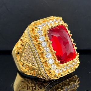 Puede incluir: Un anillo dorado con una gran gema roja rectangular como pieza central. El anillo está adornado con gemas claras más pequeñas y detalles intrincados alrededor de los bordes, creando una apariencia lujosa.