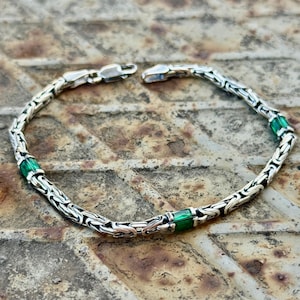 May include: A silver bracelet with a chain-like design, featuring three rectangular green gemstones. The bracelet is displayed on a textured, rusty metal surface. The clasp is open, showing the intricate details of the chain links.