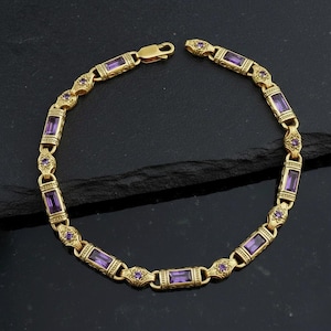 Könnte beinhalten: Goldfarbenes Armband mit rechteckigen, violetten Edelsteinen und filigranen floralen Details. Das Armband hat einen Verschluss und wird auf dunklem Untergrund gezeigt. Ein klassisches Schmuckstück.