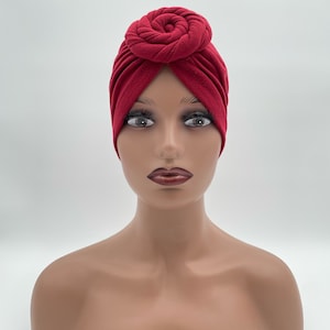 Może przedstawiać: Ciemnoczerwony turban z wyraźnym, okrągłym węzłem na środku czoła. Materiał jest miękki i matowy, z subtelnymi pionowymi plisami rozchodzącymi się od centralnego węzła, które pokrywają czubek głowy oraz jej boki, tworząc spójną całość.