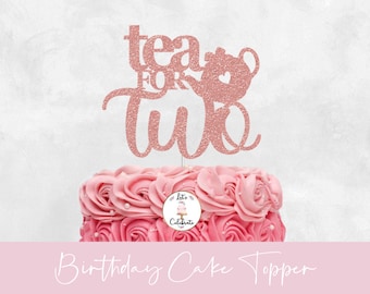 以下が含まれることがあります： ローズゴールドのグリッターケーキトッパーで、「tea for two」と書いてあり、ティーポットのシルエットが付いています。トッパーはピンクのフロスティングケーキの上に載っています。
