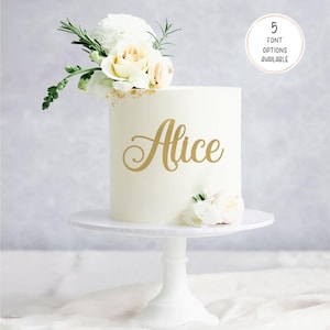 May include: A white cake decorated with the name "Alice" in gold script, and topped with white flowers and foliage. The cake is on a white stand. A small circle indicates "5 Font Options Available".