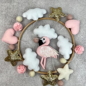May include: A pink and gold baby mobile with clouds, stars, hearts, and a flamingo. The mobile is made of felt and has a wooden hoop.