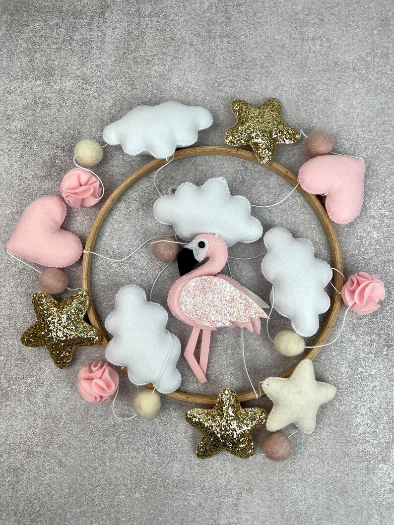 May include: A pink and gold baby mobile with clouds, stars, hearts, and a flamingo. The mobile is made of felt and has a wooden hoop.