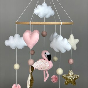 May include: A pink and white baby mobile with a pink flamingo, clouds, stars, and hearts. The mobile is made of felt and has wooden rings.