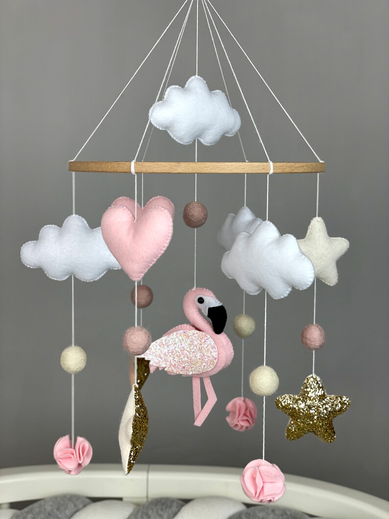 May include: A pink and white baby mobile with a pink flamingo, clouds, stars, and hearts. The mobile is made of felt and has wooden rings.