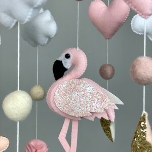 May include: A pink and white baby mobile with a pink flamingo, white clouds, pink hearts, and beige felt balls.