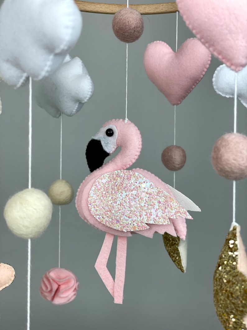 May include: A pink and white baby mobile with a pink flamingo, white clouds, pink hearts, and beige felt balls.
