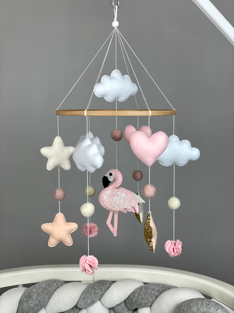 May include: A pink and white baby mobile with a pink flamingo, clouds, stars, and hearts hanging from a wooden ring.