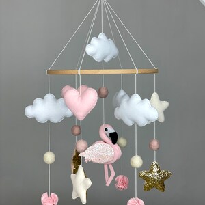 May include: A pink and white felt baby mobile with a pink flamingo, clouds, stars, and hearts. The mobile is hanging from a wooden ring.