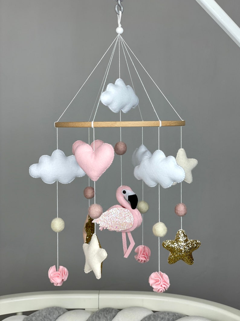 May include: A pink and white felt baby mobile with a pink flamingo, clouds, stars, and hearts. The mobile is hanging from a wooden ring.