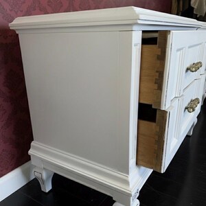 May include: White painted dresser with two visible drawers and ornate brass handles. The dresser has a classic design with decorative molding and legs. One drawer is open, revealing the wooden interior.