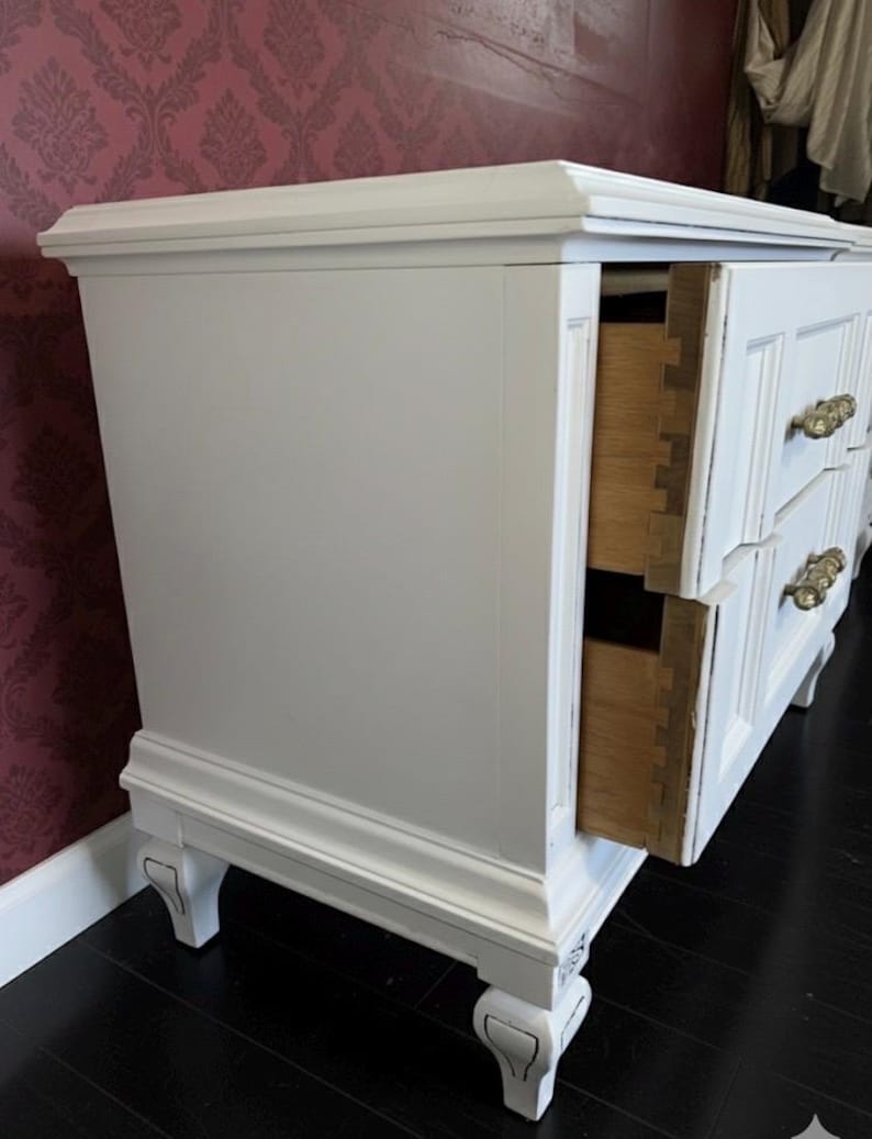 May include: White painted dresser with two visible drawers and ornate brass handles. The dresser has a classic design with decorative molding and legs. One drawer is open, revealing the wooden interior.