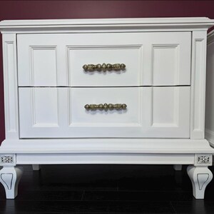 May include: White nightstand with two drawers and ornate gold-colored handles. The nightstand has decorative legs and trim. The nightstand is painted white and sits on a dark wood floor.