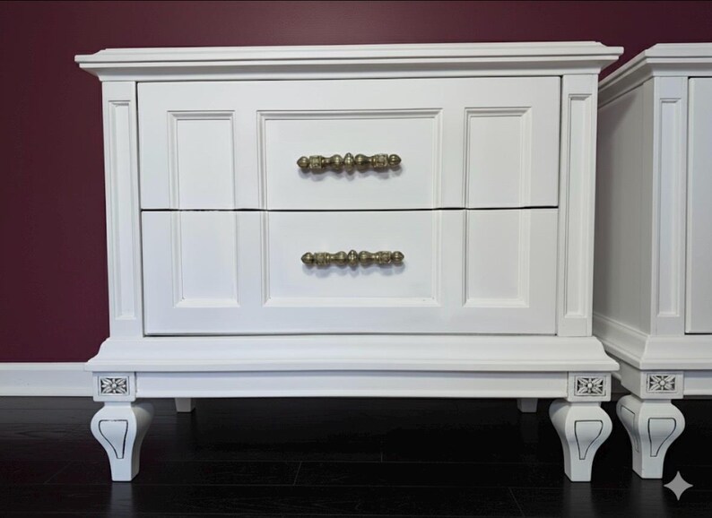 May include: White nightstand with two drawers and ornate gold-colored handles. The nightstand has decorative legs and trim. The nightstand is painted white and sits on a dark wood floor.