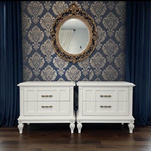 May include: Two white nightstands with two drawers each, featuring ornate hardware and legs. The nightstands are set against a backdrop of blue damask wallpaper, a gold-framed mirror, and navy blue curtains. The room has a classic, elegant aesthetic.