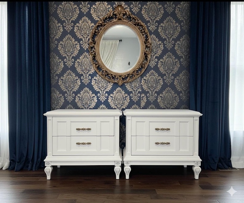 May include: Two white nightstands with two drawers each, featuring ornate hardware and legs. The nightstands are set against a backdrop of blue damask wallpaper, a gold-framed mirror, and navy blue curtains. The room has a classic, elegant aesthetic.