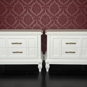 May include: Two white nightstands with two drawers each, featuring ornate brass-colored handles. The nightstands have decorative legs and are set against a burgundy damask wallpaper background.
