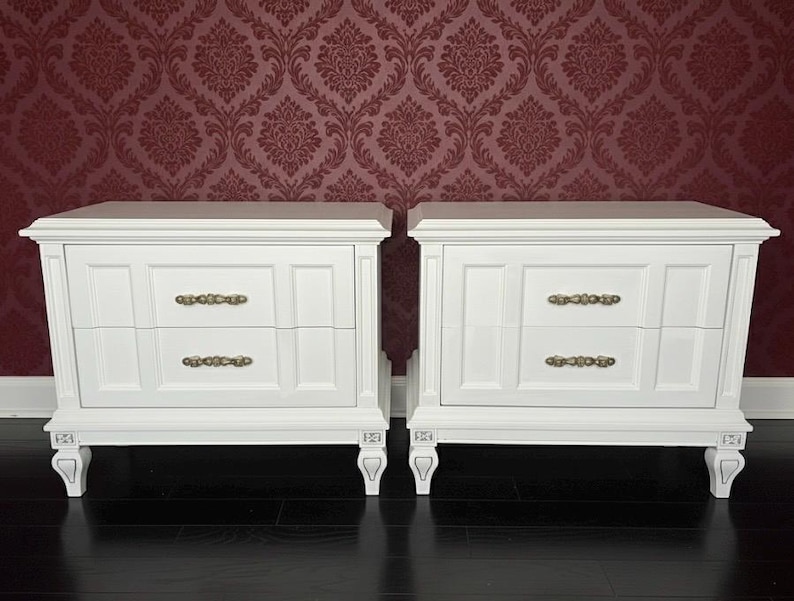 May include: Two white nightstands with two drawers each, featuring ornate brass-colored handles. The nightstands have decorative legs and are set against a burgundy damask wallpaper background.