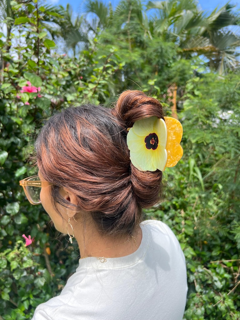 May include: A yellow and white flower hair clip is worn in a bun. The hair clip has a black center and is made of a light material. The person is wearing a white shirt and gold-rimmed glasses. The background is a lush green garden.
