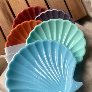 Può includere: Una collezione di piatti a forma di conchiglia in vari colori, tra cui blu, verde acqua, arancione, bordeaux e viola. I piatti sono disposti a ventaglio, mostrando i loro bordi smerlati e le superfici strutturate. Lo sfondo comprende una doga di legno e un tessuto neutro.