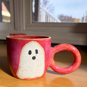 May include: A ceramic mug with a vibrant pink speckled glaze featuring a hand-painted white ghost shape on the front. The ghost has three simple black dots for eyes and a mouth. The mug includes a large, thick, circular handle on the right side.