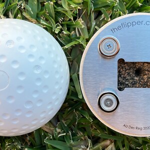 K&ouml;nnte beinhalten: Ein wei&szlig;er, mit Noppen besetzter Ball mit flachem Boden und einer silbernen Metallbasis mit vier Magneten. Die Basis tr&auml;gt den Text "theflipper.com.au" und "AU Des Reg 2020/4958".