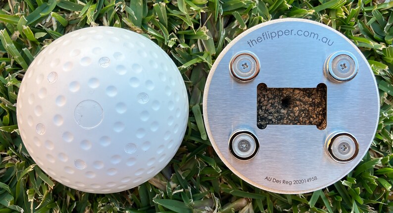 K&ouml;nnte beinhalten: Ein wei&szlig;er, mit Noppen besetzter Ball mit flachem Boden und einer silbernen Metallbasis mit vier Magneten. Die Basis tr&auml;gt den Text "theflipper.com.au" und "AU Des Reg 2020/4958".