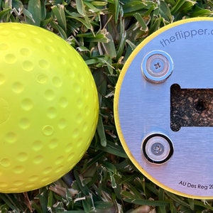 K&ouml;nnte beinhalten: Ein gelber Golfball mit Dimples und einem silbernen Metallsockel mit vier Magneten und dem Text "theflipper.com.au" und "AU Des Reg 202014958".