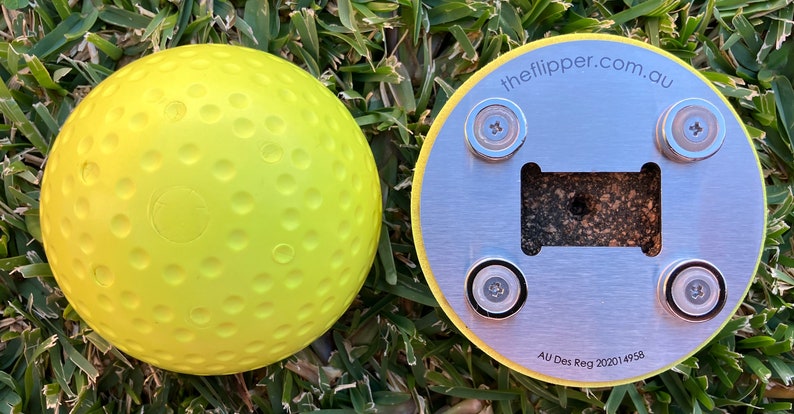K&ouml;nnte beinhalten: Ein gelber Golfball mit Dimples und einem silbernen Metallsockel mit vier Magneten und dem Text "theflipper.com.au" und "AU Des Reg 202014958".
