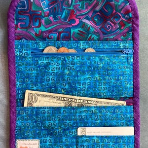 May include: A fabric wallet with a purple border and a blue interior featuring a geometric square pattern. It includes a zippered coin pocket, a bill compartment containing a one-dollar bill, a card slot with a card, and a label reading Handmade by Joan.
