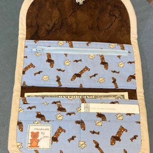 May include: A fabric wallet with a light blue interior featuring a repeating pattern of brown dachshunds and yellow houses. The wallet includes a zippered compartment, card slots, a bill pocket, a snap closure, and a label reading Handmade by Joan.