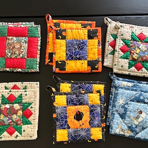 May include: Six sets of square quilted pot holders featuring various seasonal patterns. Designs include deer in green frames, orange and black spiderwebs, holiday stars with red and green accents, and blue fabric with snowman and snowflake motifs.