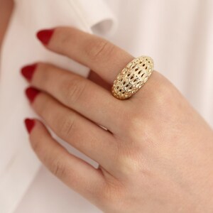 Può includere: Un anello dorato con un design ovale traforato. L'anello è indossato su un dito, su sfondo di camicia bianca. Il motivo intricato dell'anello è visibile, mostrando la sua dettagliata maestria artigianale.