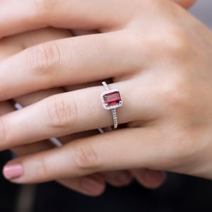 Może przedstawiać: Srebrny pierścionek z czerwonym kamieniem szlachetnym otoczonym mniejszymi diamentami.