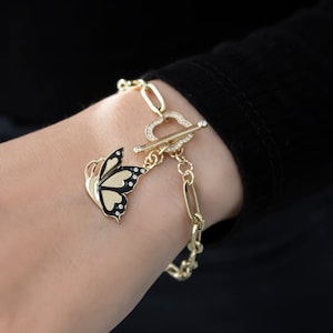 K&ouml;nnte beinhalten: Ein goldenes Kettenarmband mit einem Schmetterlingsanh&auml;nger. Das Armband hat einen herzf&ouml;rmigen Verschluss mit klaren Strasssteinen. Der Schmetterlingsanh&auml;nger ist schwarz und gold mit klaren Strasssteinen.