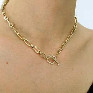Op de afbeelding: Een gouden ketting met een groot, ovaalvormig schakellinkontwerp. De ketting heeft een toggle-sluiting met een diamantbezette bloememotief.
