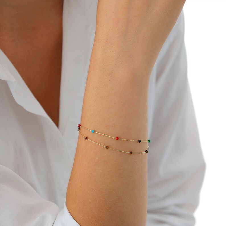 May include: Two gold chain bracelets with small colorful beads. The top bracelet has red, turquoise, and black beads. The bottom bracelet has brown beads.
