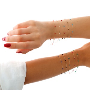 May include: A set of gold bracelets with small coloured beads. The bracelets are layered on a person's wrist. The beads are red, white, blue, green, and black.