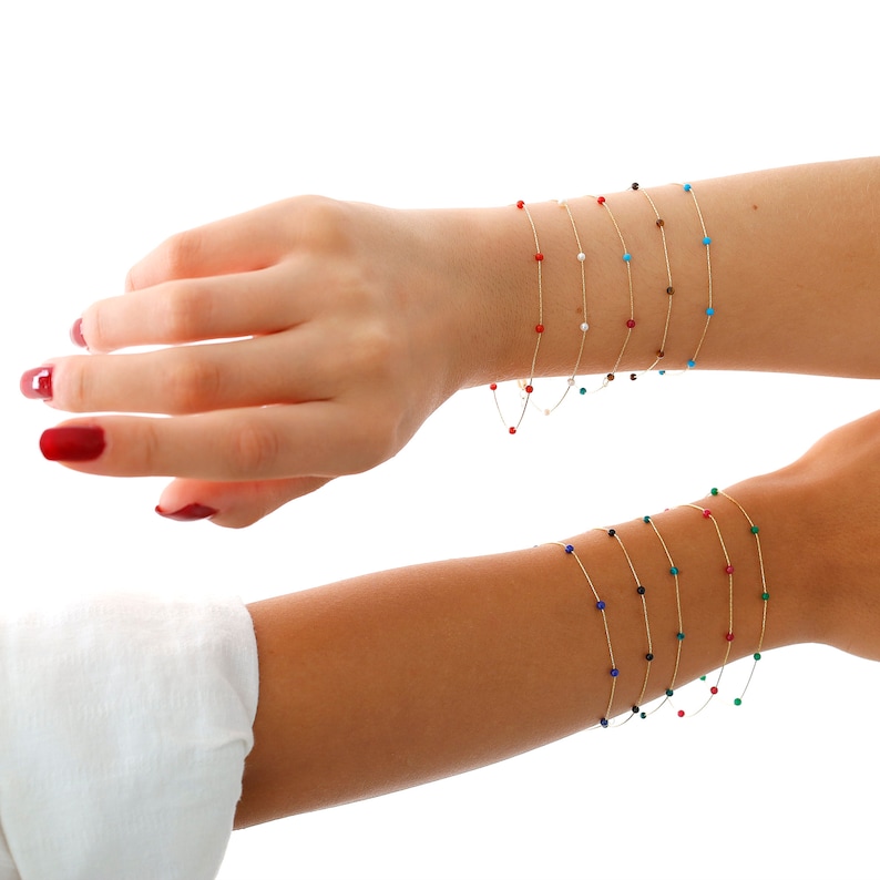 May include: A set of gold bracelets with small coloured beads. The bracelets are layered on a person's wrist. The beads are red, white, blue, green, and black.