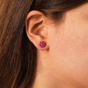 May include: Close-up of a person's ear wearing a pair of ruby stud earrings. The earrings are set in gold and feature a cluster of small, red rubies.