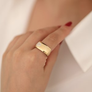 Può includere: Un anello d'oro con un design a forma di cuore di piccole pietre trasparenti sulla parte superiore. L'anello è largo e ha una superficie liscia e arrotondata.